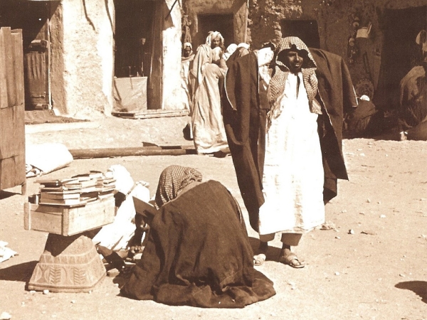 Historical picture of a person selling books In the middle of the market while browsing a book. King Abdulaziz Foundation for Research and Archives (Darah)